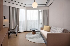 Habitación de hotel moderna con una mesa redonda, un sofá, una silla y un ventanal con cortinas.