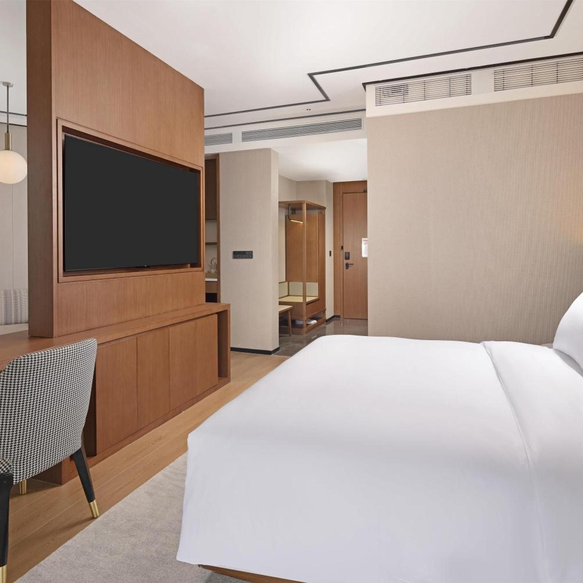 A hotel room with a large bed, a flat-screen TV mounted on a wooden panel, a houndstooth-patterned chair, and a door leading to a bathroom.