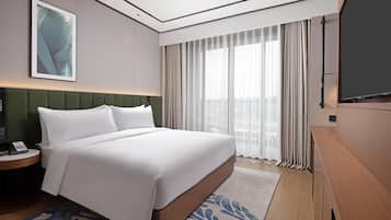A hotel room with a large bed, a green headboard, a bedside table with a phone, a large window with curtains, and a flat-screen TV.