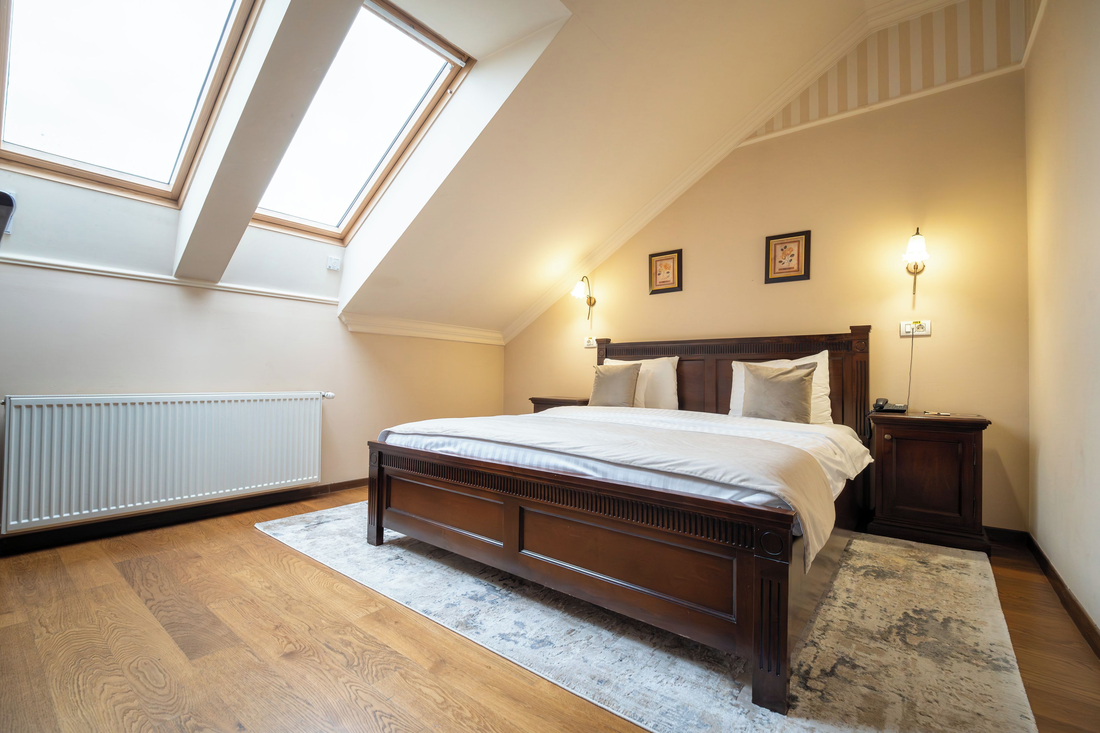 A bedroom with a sloped ceiling, a large bed, two bedside tables, a radiator, and a skylight.