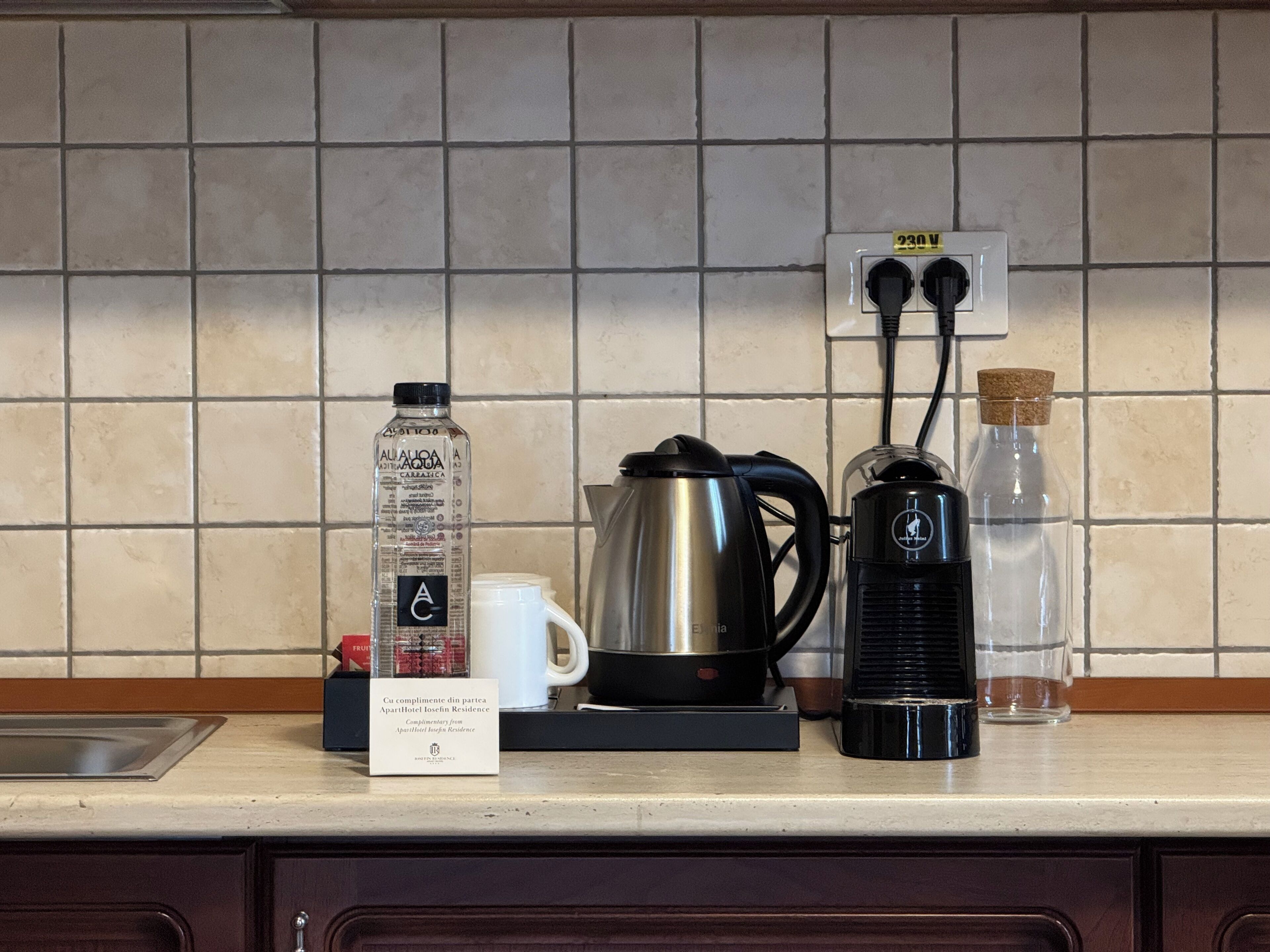 A kitchen counter with a coffee maker, a kettle, a bottle of water, a mug, and a sign.