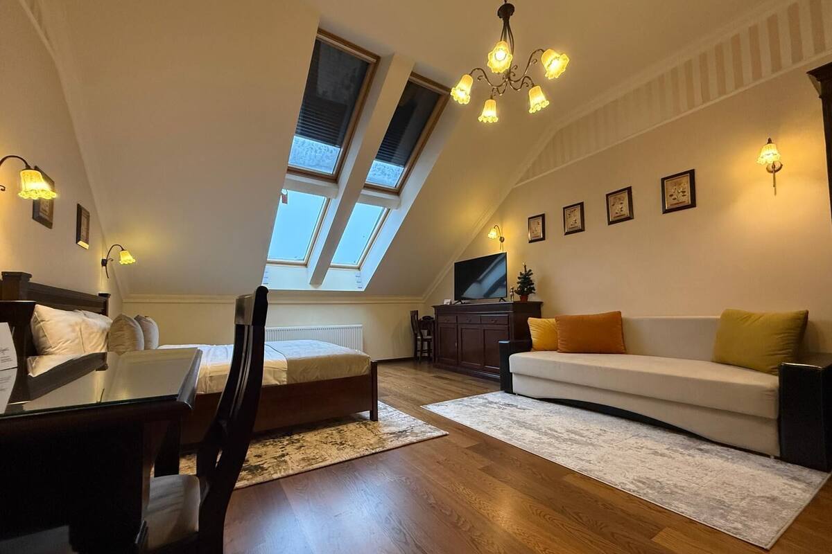 A cozy loft-style bedroom with a bed, a desk, and a sofa. There are skylights, a chandelier, and framed pictures on the walls.