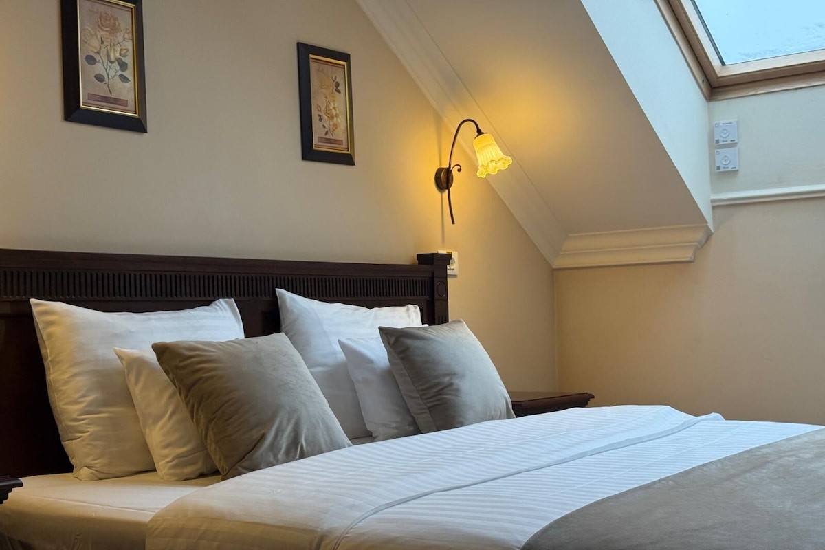 Una cama bien tendida con ropa de cama blanca y gris, un cabecero de madera y una lámpara fijada a la pared.