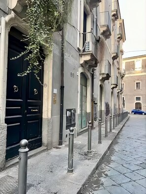A street with a row of buildings featuring dark doors and metal bollards. - Appartamento 150 mq. per famiglie e gruppi di amici (Catania)