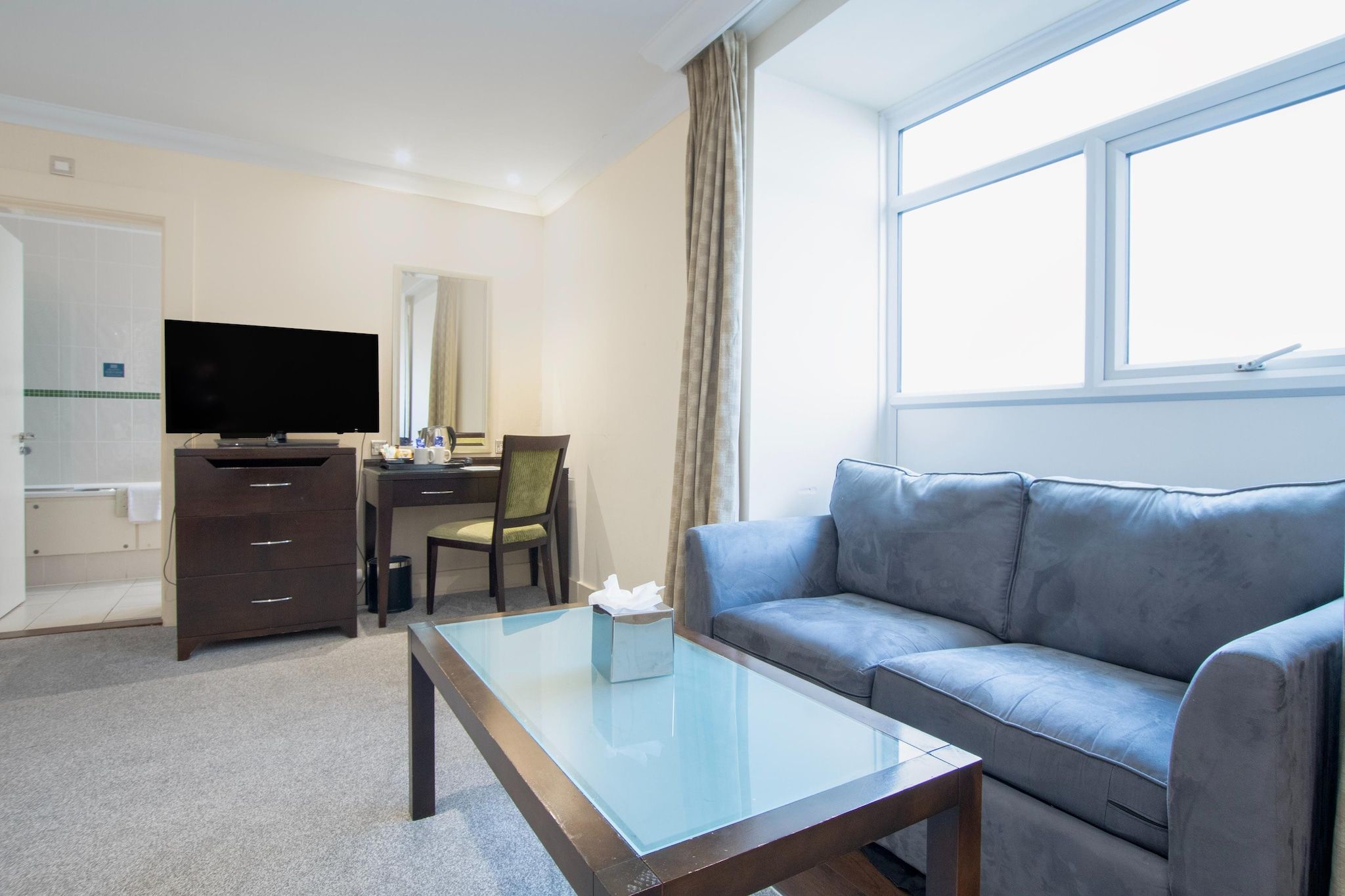 Habitación de hotel con sofá, una mesa de centro de vidrio, un escritorio con silla, televisor y ventana con cortinas.