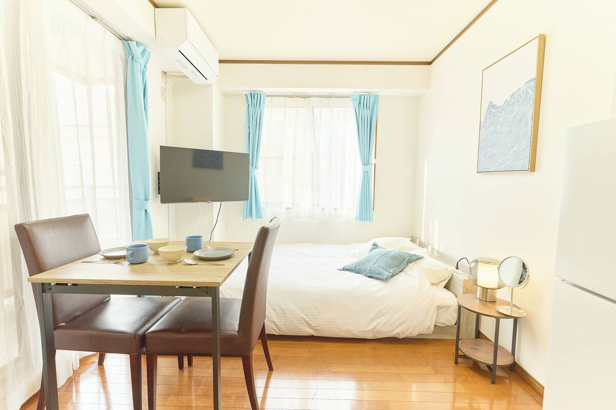 A room with a bed, a desk with chairs, a TV, a bedside table with a lamp, and a painting on the wall.