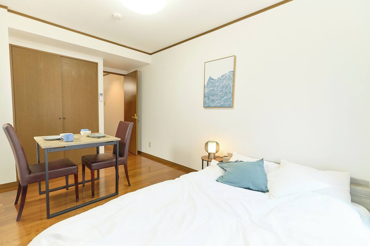 A bedroom with a bed, a bedside table with a lamp, a desk with chairs, and a door leading to another room.