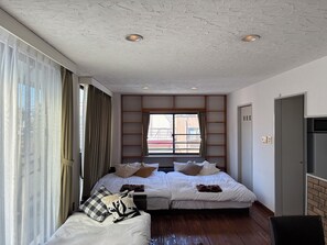 Quarto de hotel com duas camas, uma janela grande com cortinas, uma prateleira embutida e uma porta.