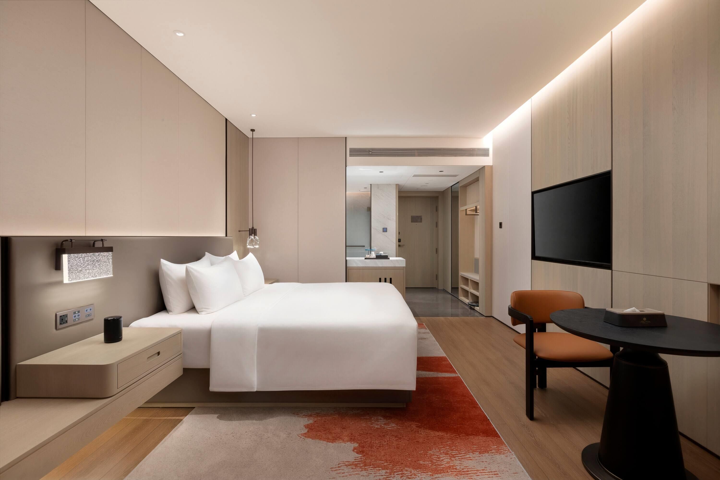 A modern hotel room with a bed, bedside table, lamp, TV, and a small table with a chair.