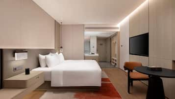 A modern hotel room with a bed, bedside table, lamp, TV, and a small table with a chair.