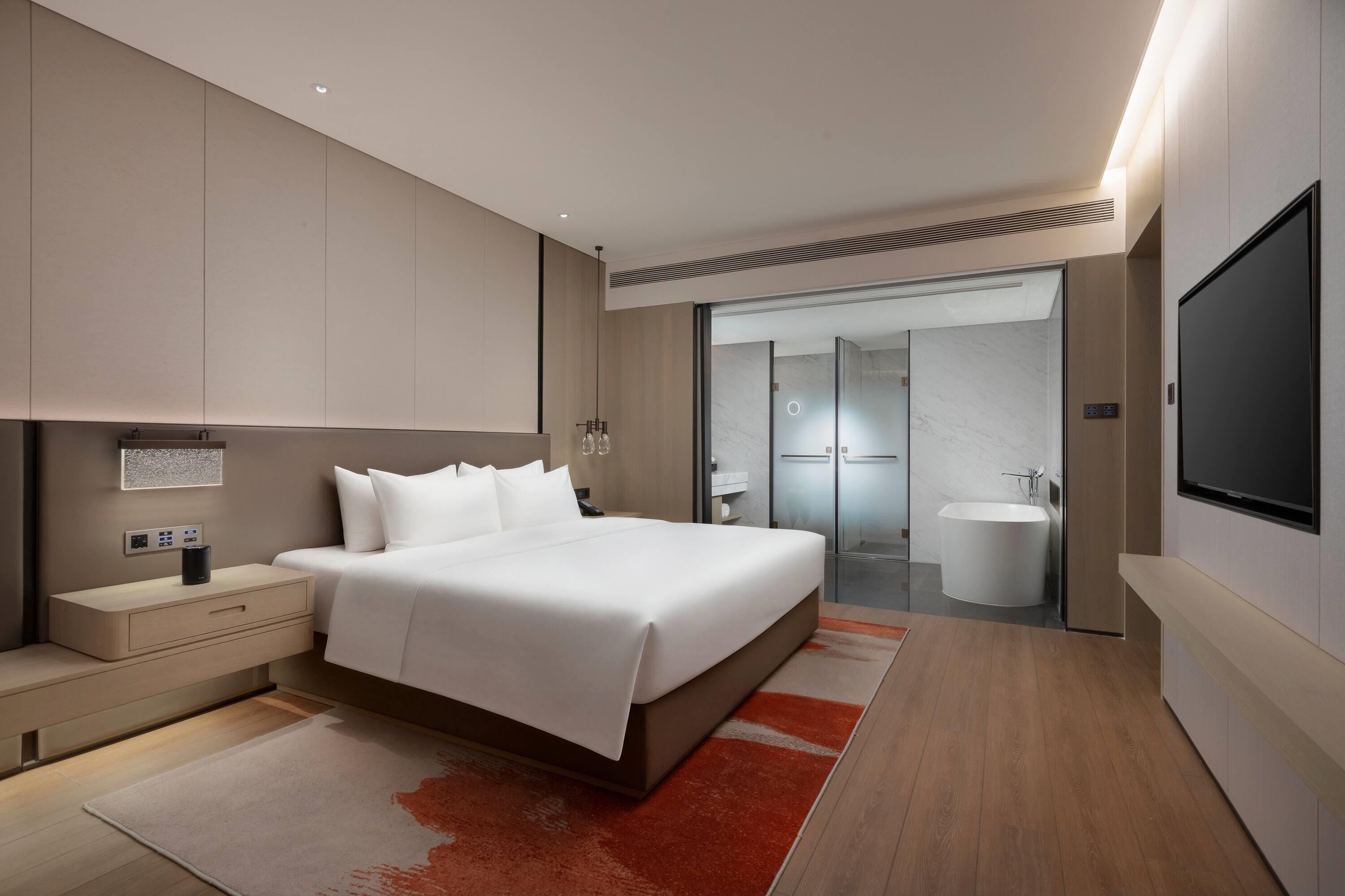 A modern hotel room with a large bed, bedside tables, a flat-screen TV, a bathroom with a bathtub, and a shower.
