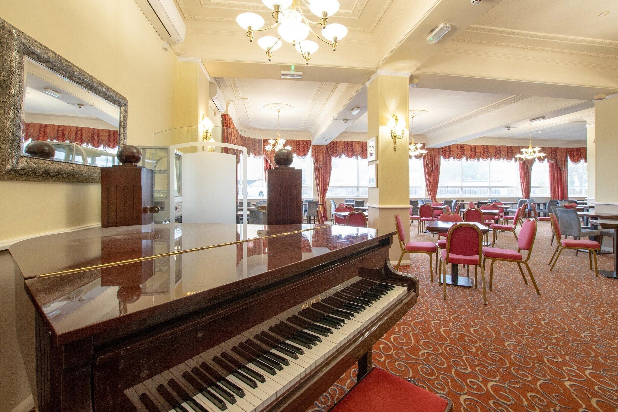 Un piano à queue dans la salle à manger d’un hôtel, avec des chaises à revêtement rouge et un tapis à motifs.