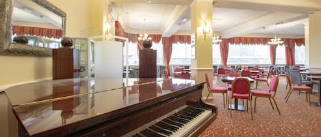 A grand piano in a hotel dining area with red upholstered chairs and a patterned carpet.