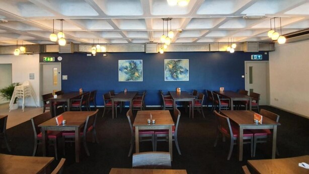 A restaurant with multiple tables and chairs, ceiling lights, and two paintings on the wall.