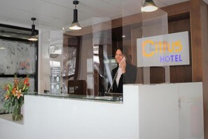 A hotel reception desk with a glass partition, a clock, and a sign for 'Citus Hotel'.