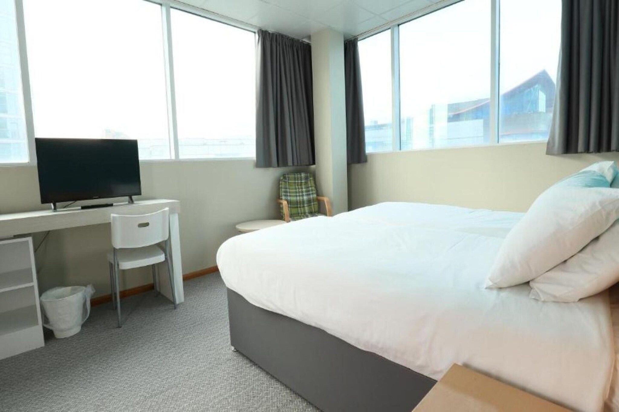 A hotel room with a bed, a desk with a chair, a television, and large windows.