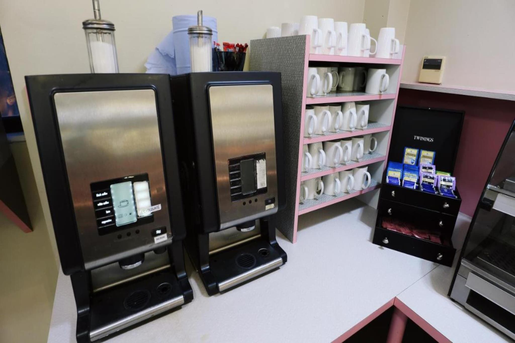 Zwei Espressomaschinen mit digitalen Anzeigen, ein Regal mit weißen Tassen und eine Vitrine mit Twinings-Teesorten.