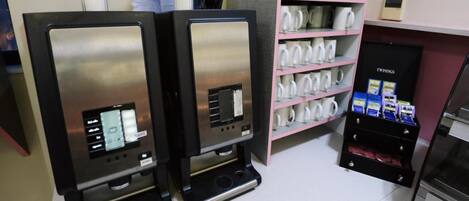 Zwei Espressomaschinen mit digitalen Anzeigen, ein Regal mit weißen Tassen und eine Vitrine mit Twinings-Teesorten.