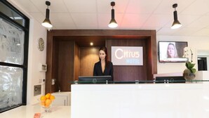 Una mesa de recepción en el Hotel Citrus, con una pantalla digital que muestra el nombre del hotel, un reloj y un jarrón con flores.