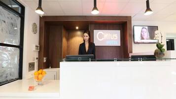 A reception desk at the Citrus Hotel with a digital screen displaying the hotel name, a clock, and a vase with flowers.