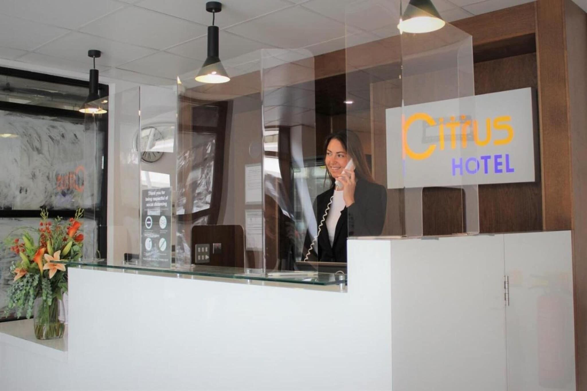 A hotel reception desk with a glass partition, a clock, and a sign for 'Citus Hotel'.