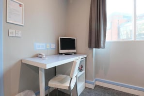 A small office space with a desk, chair, and a computer monitor. There is a window with curtains, a wall-mounted telephone, and a framed notice. - Standard Triple Room (Cardiff)