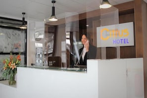 Un mostrador de recepción de hotel con una mampara de vidrio, un reloj y un letrero que dice 'Citus Hotel'.