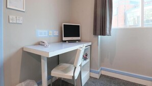 Un pequeño espacio de oficina con escritorio, silla y monitor de computadora. Hay una ventana con cortinas, un teléfono de pared y un aviso enmarcado.