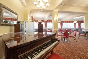 Un piano à queue dans la salle à manger d’un hôtel, avec des chaises à revêtement rouge et un tapis à motifs.