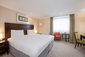 Quarto de hotel com uma cama grande, uma escrivaninha, uma cadeira, uma luminária e uma janela com cortinas.