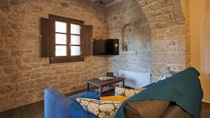 Ein Zimmer mit Steinwänden und einem Fenster mit Holzrahmen, einem an der Wand befestigten Flachbildfernseher, einem kleinen Tisch mit einer Schüssel und einer Pflanze sowie einem Sofa mit gemusterten Kissen.