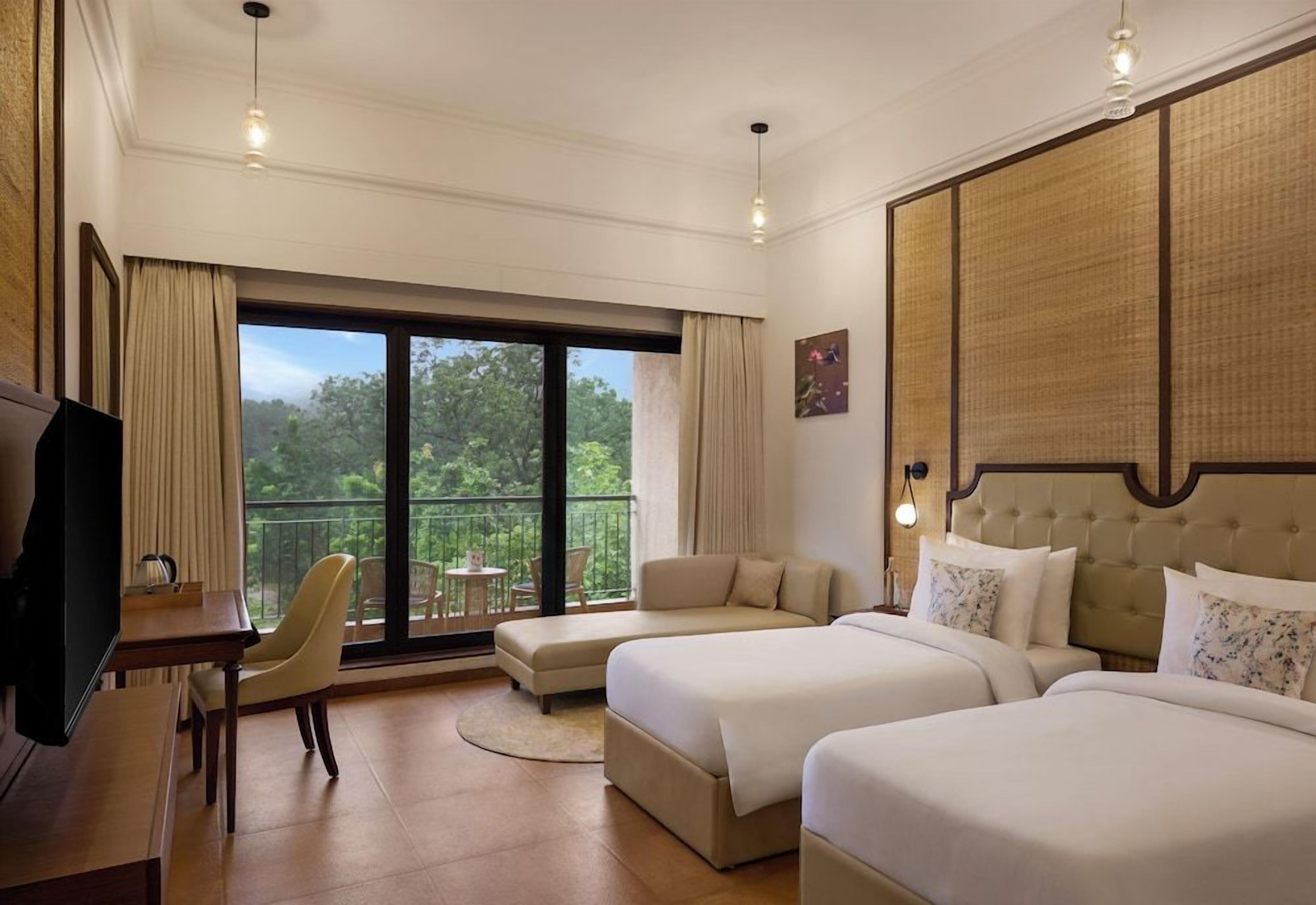 Habitación de hotel con dos camas, un escritorio, una silla y un balcón con vistas a árboles.