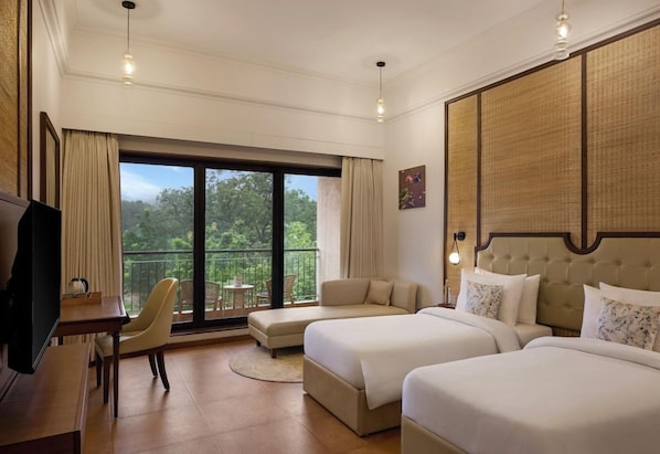 A hotel room with two beds, a desk, a chair, and a balcony with a view of trees.