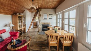 A cozy living room with a wooden ceiling, a fireplace, a dining area with wooden chairs, and a red sofa.