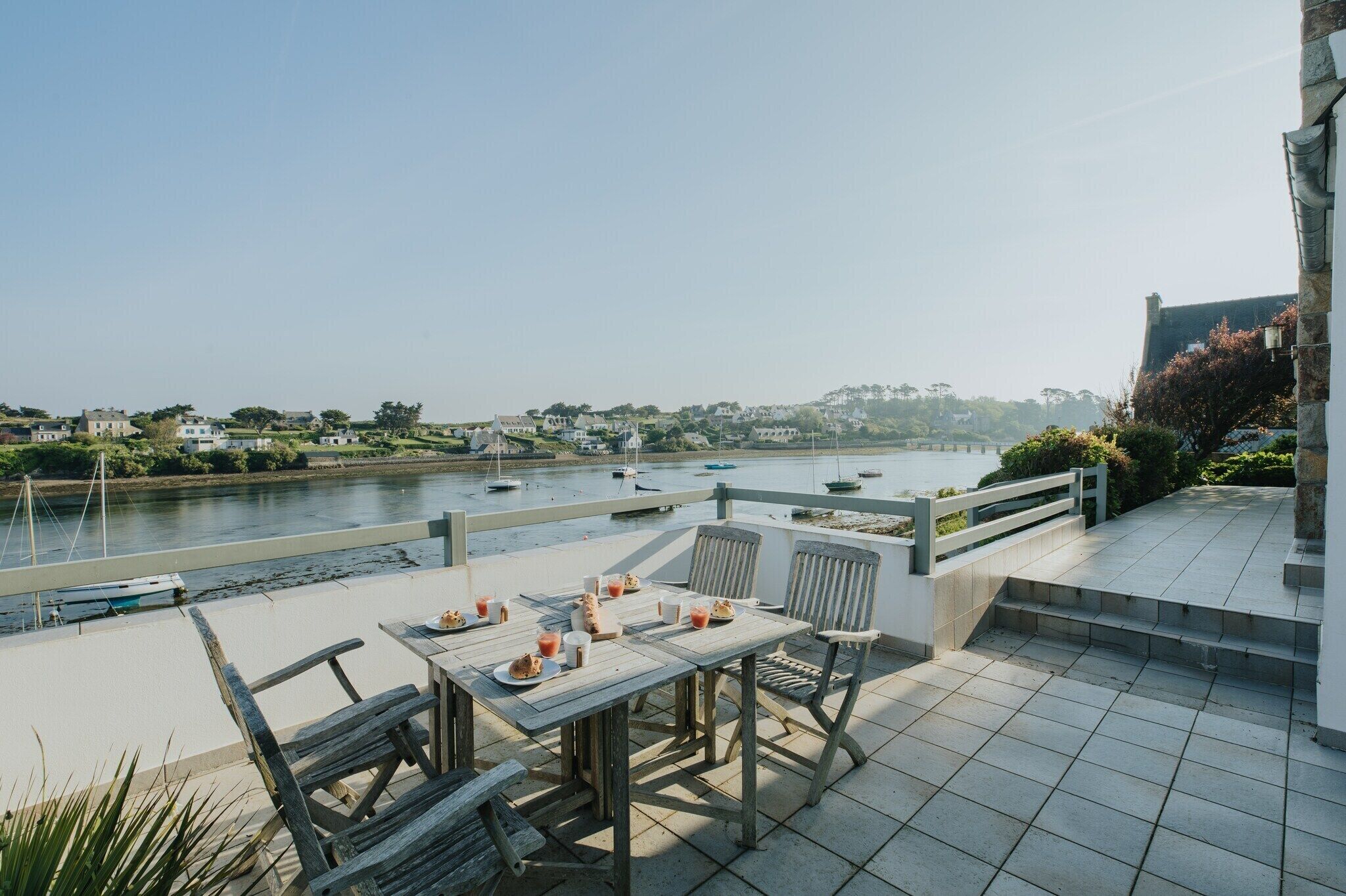 A terrace with a table set for dining, overlooking a river with boats.