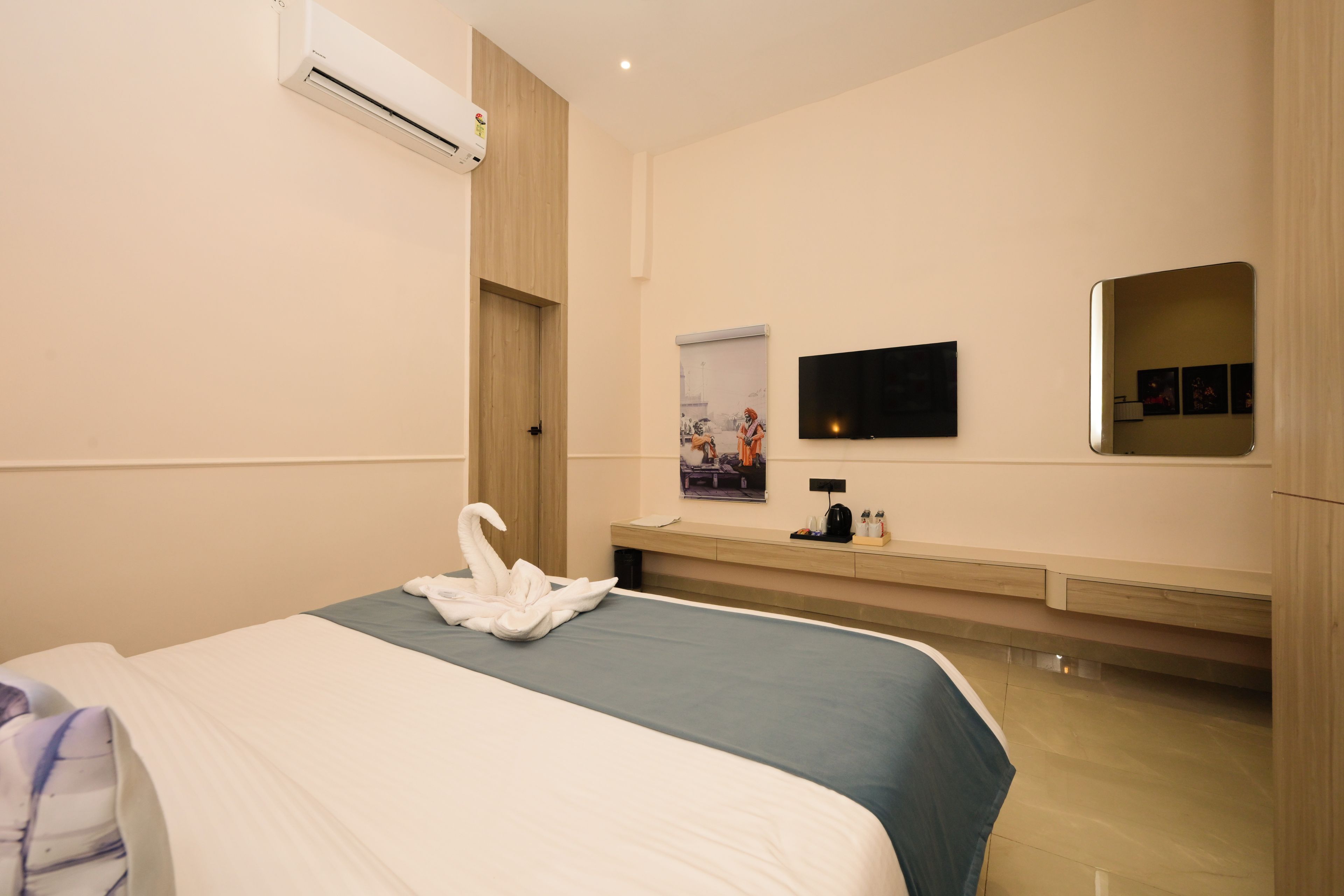 A hotel room with a bed, a flat-screen TV mounted on the wall, a wooden door, and a small table with a kettle and cups.