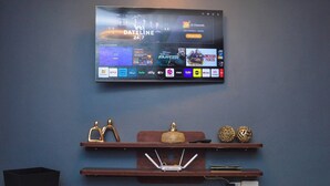 Un televisor de pantalla plana montado en la pared muestra una interfaz de servicio de transmisión con varias opciones de canales. Debajo del televisor, hay repisas de madera con objetos decorativos y una regleta de enchufes.