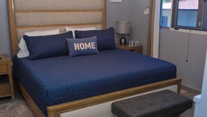 Una cama bien hecha con colcha y almohadas azules, cabecera de madera, mesita de noche con lámpara y un banco al pie de la cama.