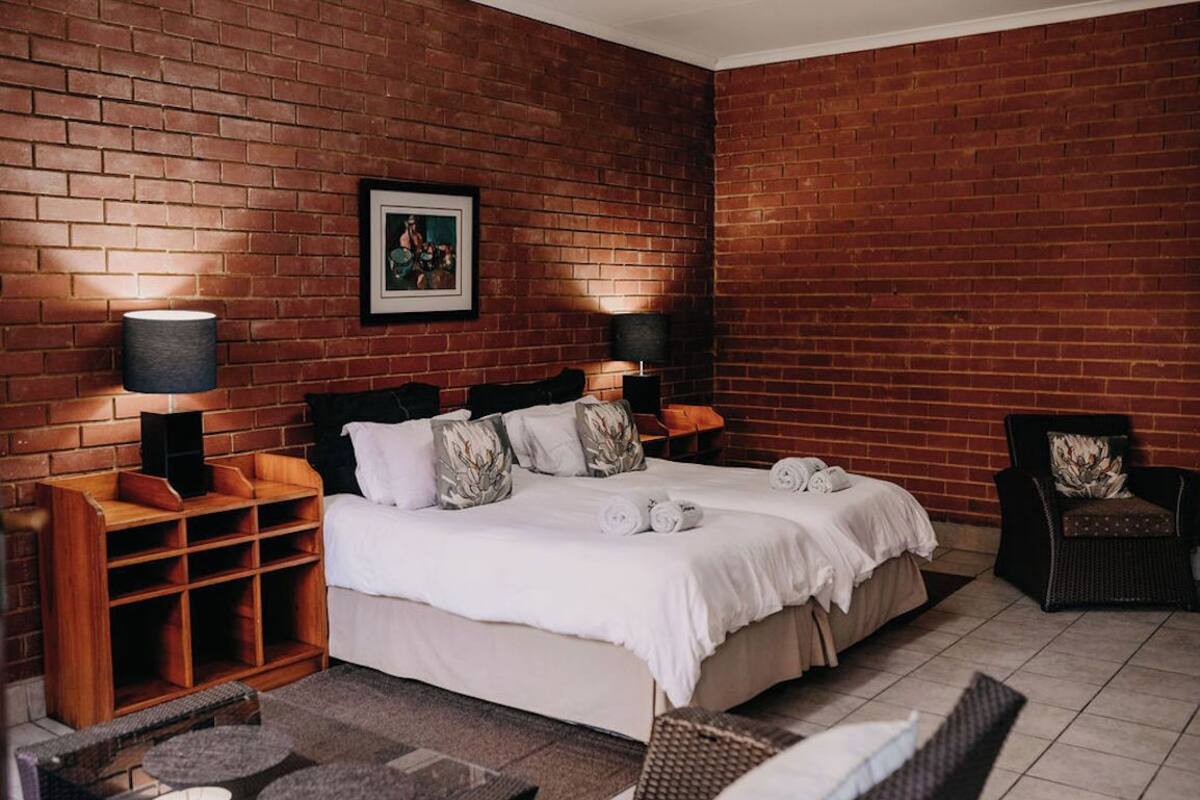 A bedroom with a brick wall, a bed with white linens, two bedside tables with lamps, a wooden side table, and a wicker chair.