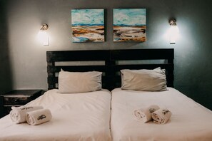 A double bed with white linens and two pillows, a nightstand with a lamp, and two paintings on the wall.