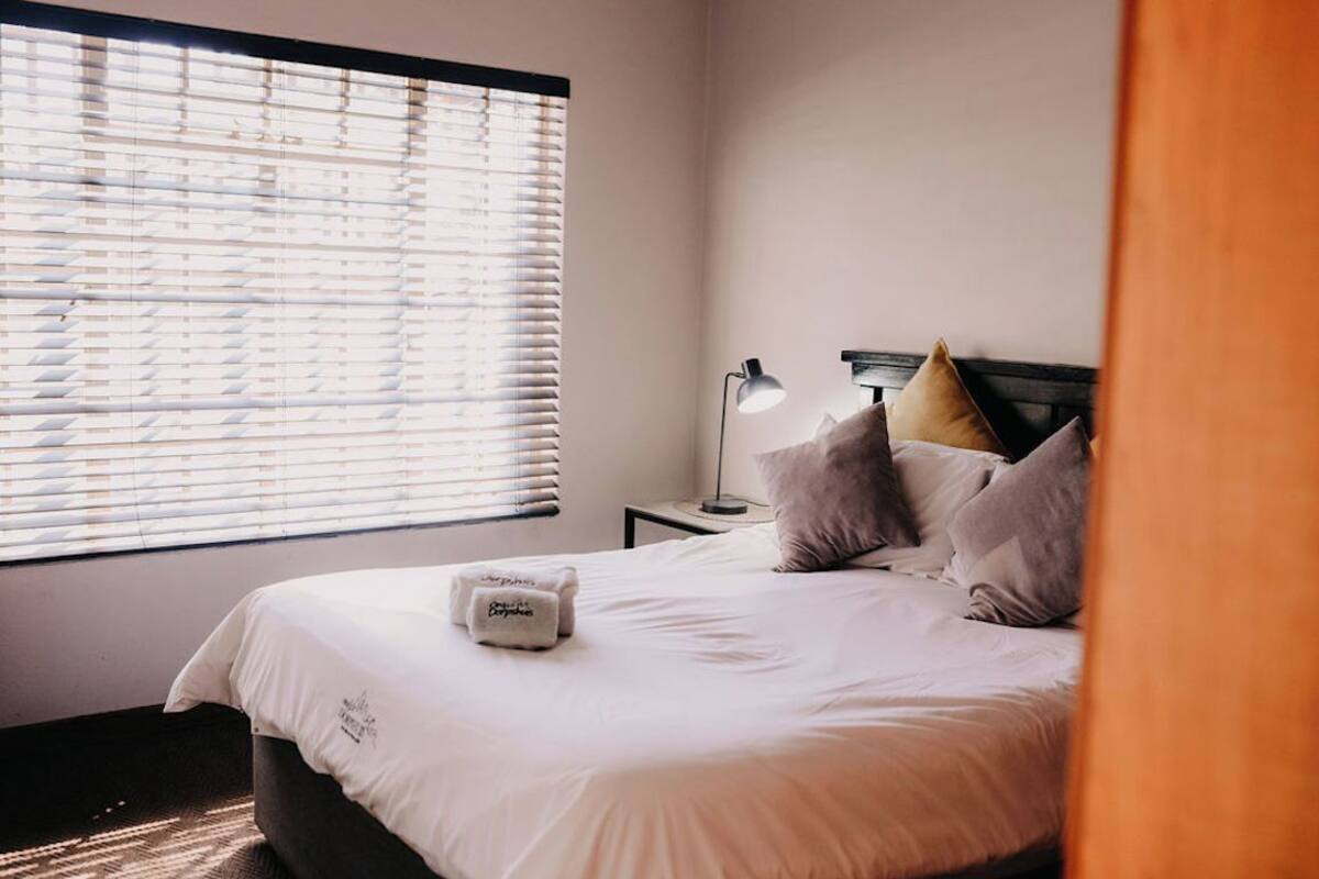 A neatly made bed with pillows and a blanket, a bedside lamp, and a window with blinds.