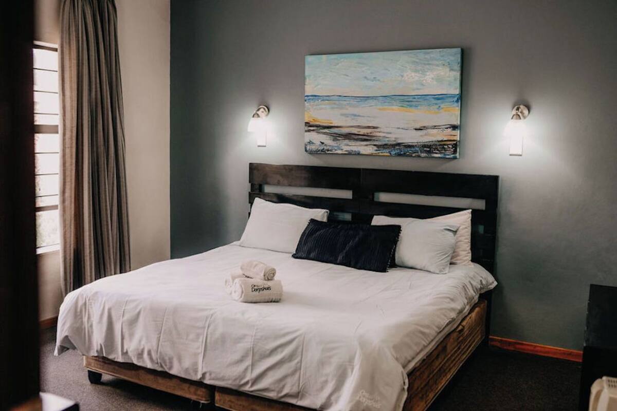 A neatly made bed with white linens and a folded towel, a painting of a beach scene above it, and two wall-mounted lights on either side.