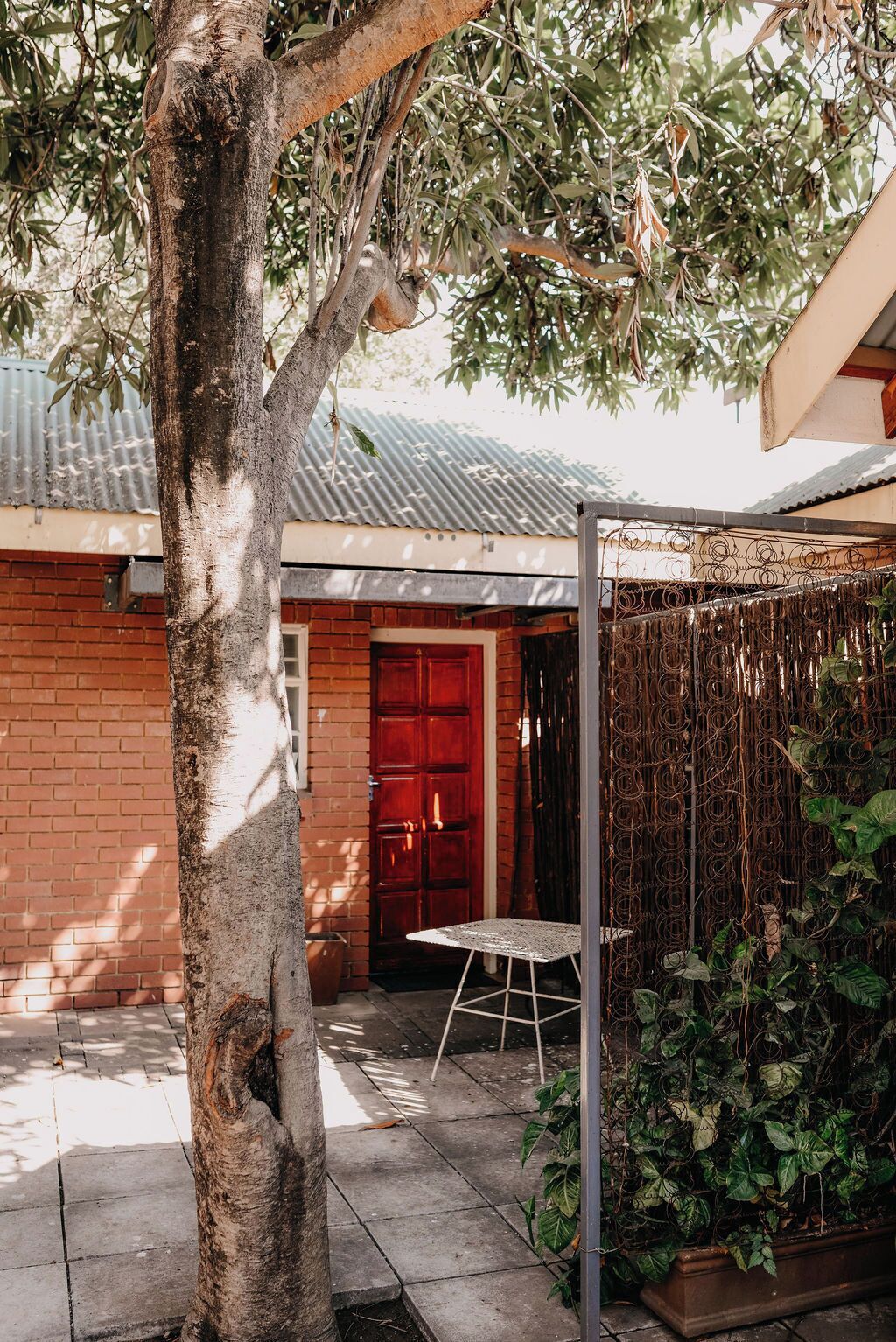 Una casa de ladrillo con una puerta roja, una mesa metálica y un árbol en primer plano.