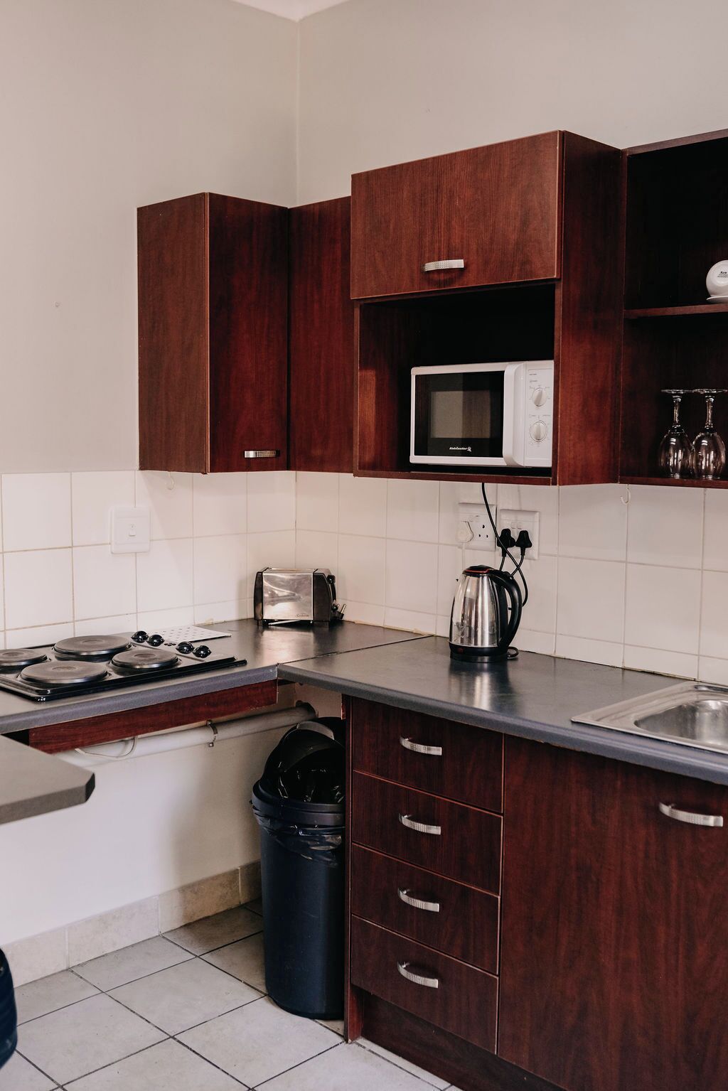 A kitchen with dark wooden cabinets, a microwave, a toaster, a kettle, and a trash bin.