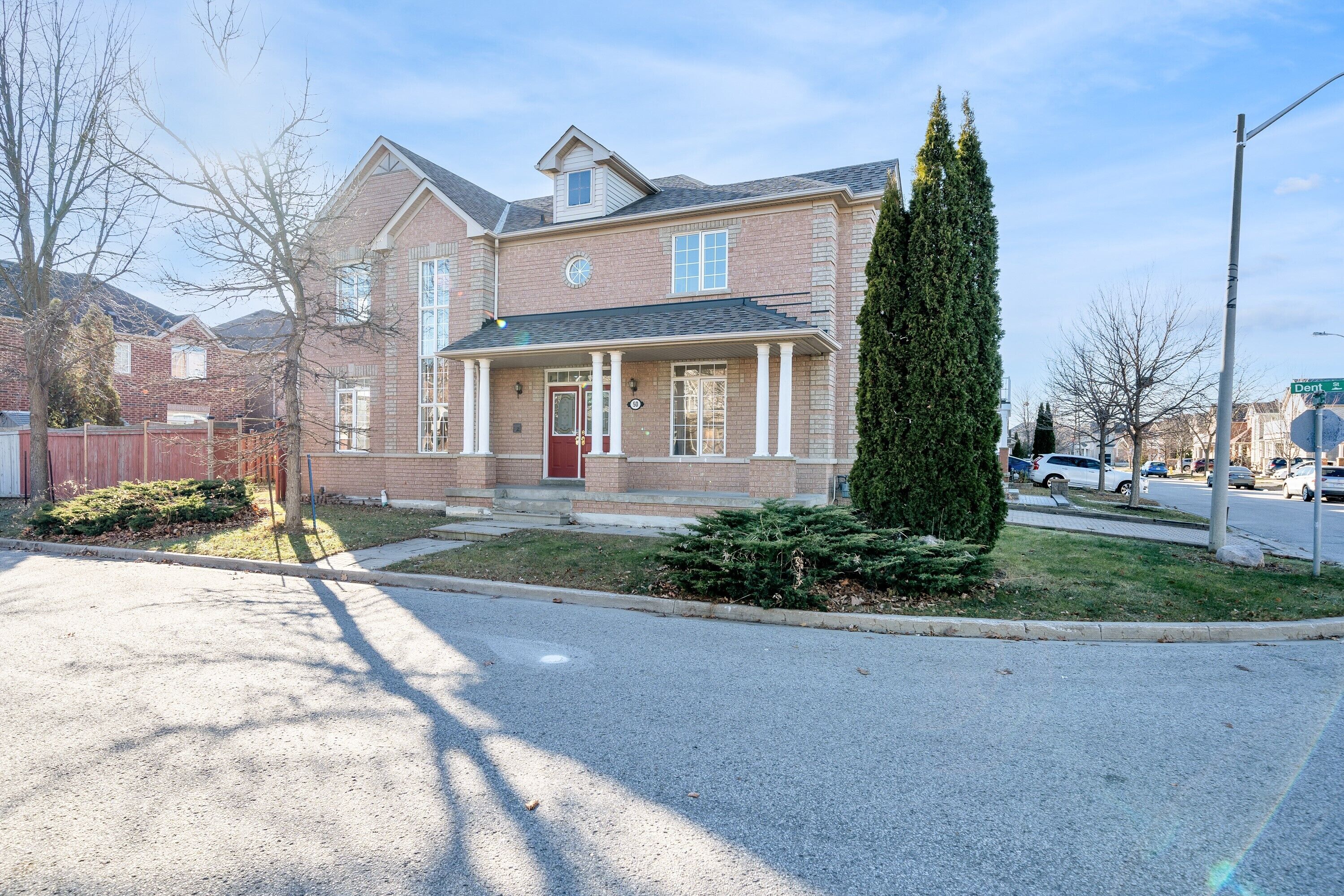 A two-story brick house with a front porch, a tall evergreen tree, and a street view.