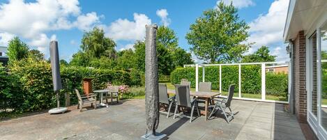 An outdoor patio with a table and chairs, surrounded by greenery and a clear sky.