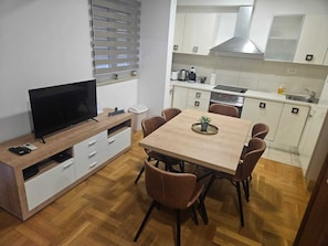 Un espacio habitable compacto con televisor, mesa de comedor, sillas, una pequeña cocina y un ventanal con persianas.