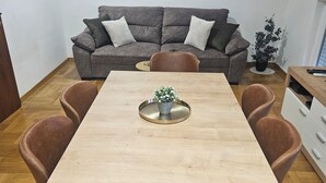 Et spiseområde med et træbord, seks læderstole i brunt, en grå sofa med to hvide puder, et billede på væggen og en potteplante.