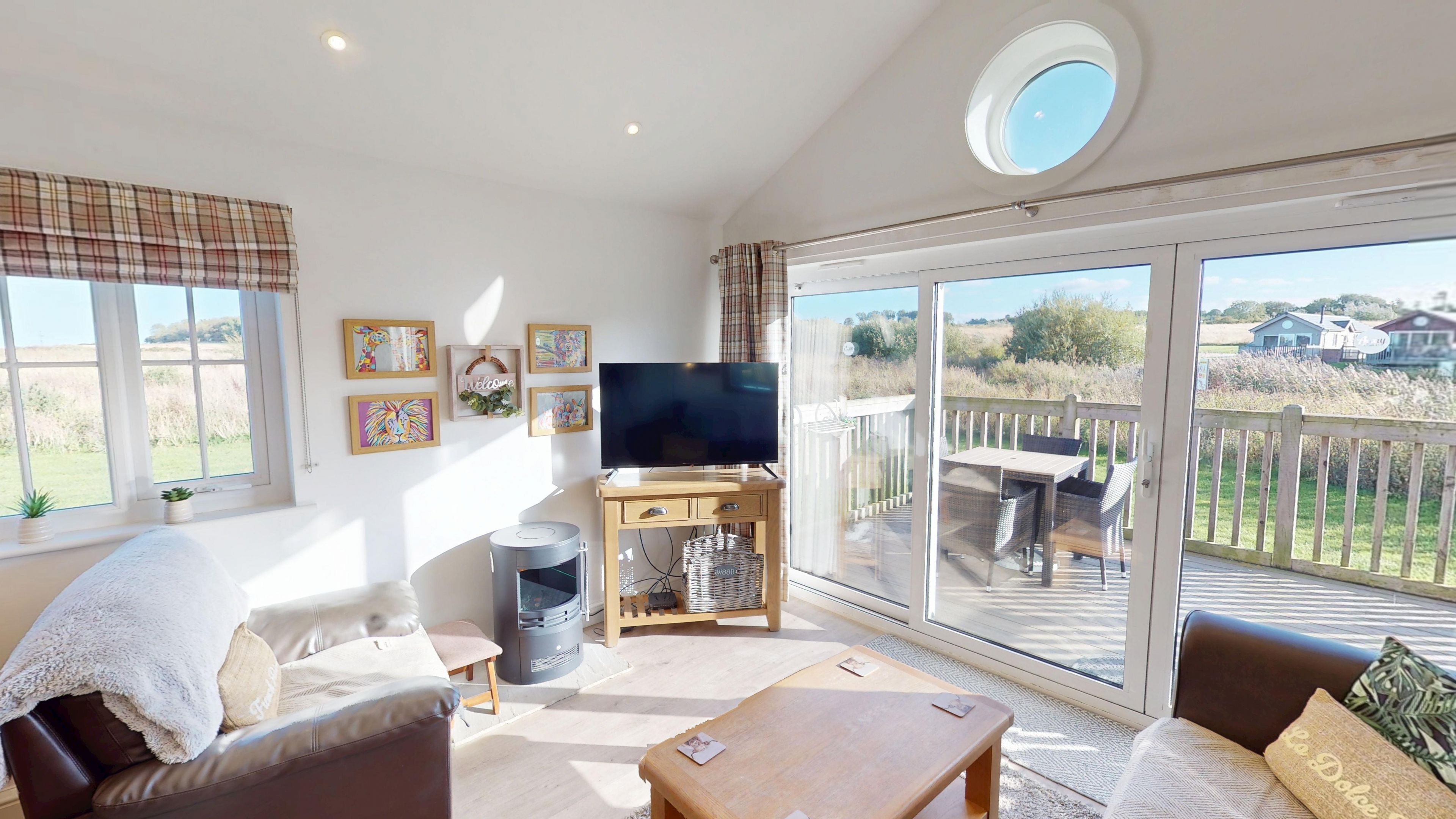 A spacious living room with a flat-screen TV, a sofa, a coffee table, and a fireplace. There are large windows offering a view of the outdoors.