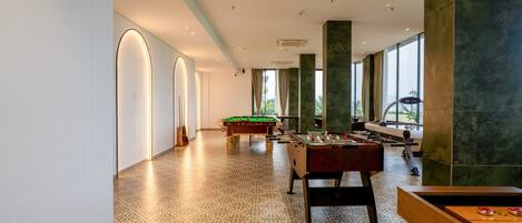 A spacious indoor area with a pool table, foosball table, and a billiards table, featuring a patterned floor and large windows.